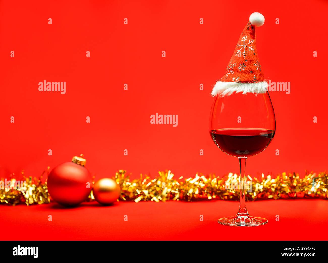 Un verre de vin rouge surmonté d'un chapeau de Père Noël, placé sur une surface rouge vif avec des ornements de Noël et des guirlandes dorées en arrière-plan, évoquant un fès Banque D'Images