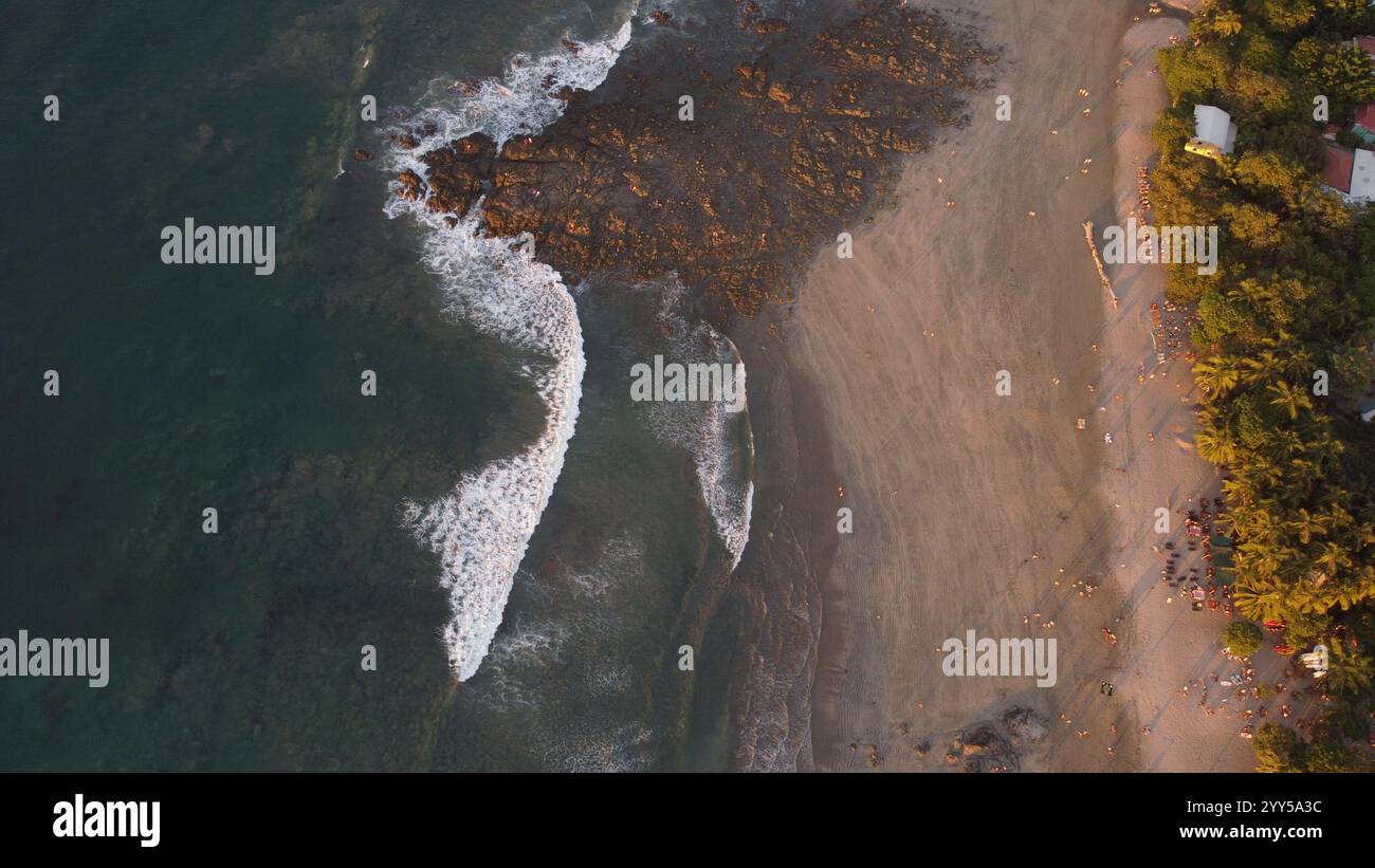 Vue aérienne d'une plage tropicale au coucher du soleil avec des palmiers, rivage rocheux, et les visiteurs dispersés appréciant le paysage côtier serein. Banque D'Images