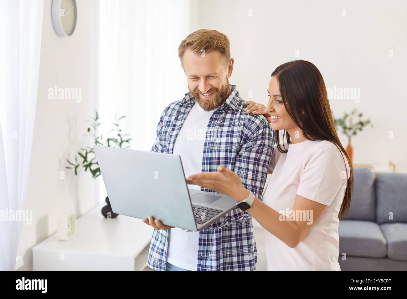 Jeune couple heureux debout dans un salon confortable regardant l'écran d'ordinateur portable, regardant la vidéo, achetant Banque D'Images