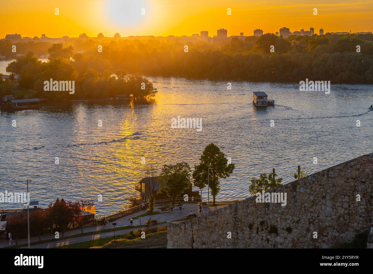 Le soleil se couche sur la rivière Sava, projetant des teintes dorées sur l'eau. Depuis la forteresse de Kalamegdan à Belgrade, la vue met en valeur la nature paisible et l'horizon de la ville se mélangeant magnifiquement. Banque D'Images