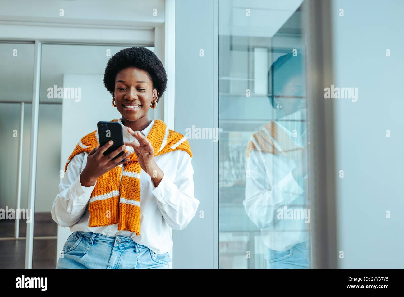 Une femme architecte confiante dans une chemise blanche et un pull orange, debout dans un environnement de bureau élégant, s'engageant avec son smartphone. Son exp joyeux Banque D'Images