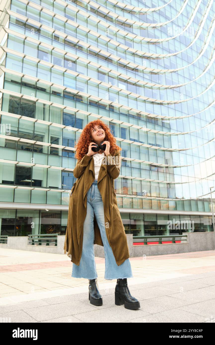 Jeune femme élégante portant des écouteurs regardant vers le bâtiment moderne Banque D'Images