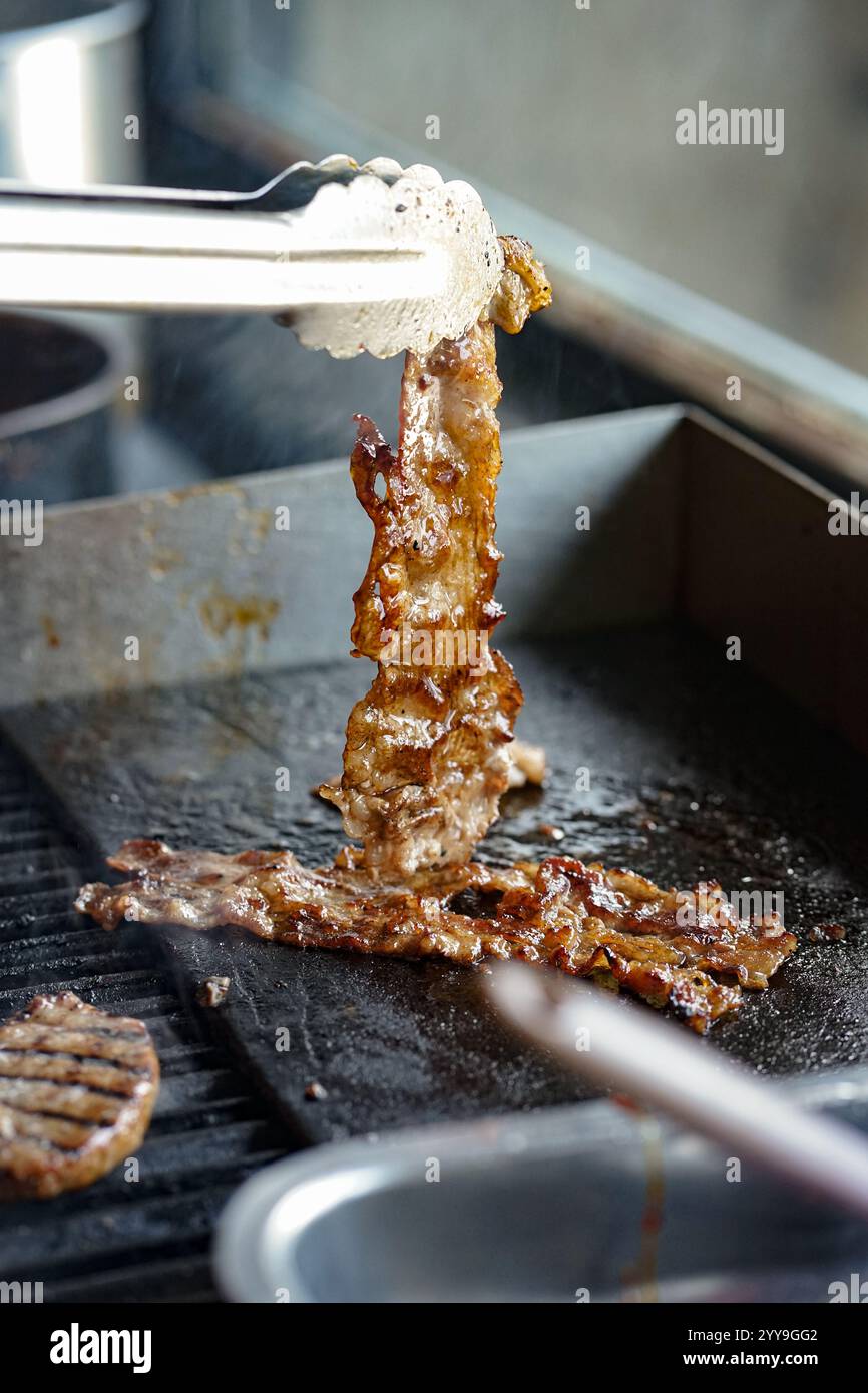 Gros plan de bœuf grillé coréen grillé sorti d'un gril plat chaud à l'aide d'une pince métallique, étincelant de jus et carbonisé à la perfection. Banque D'Images