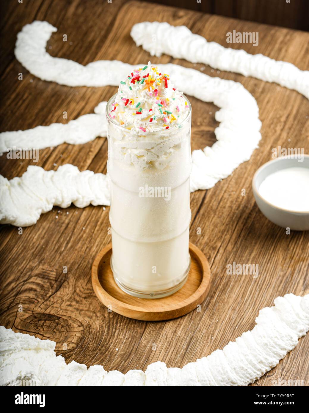 Un grand milk-shake à la vanille surmonté de crème fouettée et de pépites colorées, stylisé de manière créative avec des tourbillons de crème fouettée sur une surface en bois rustique Banque D'Images Un grand milk-shake à la vanille surmonté de crème fouettée et de pépites colorées, stylisé de manière créative avec des tourbillons de crème fouettée sur une surface en bois rustique Banque D'Images
