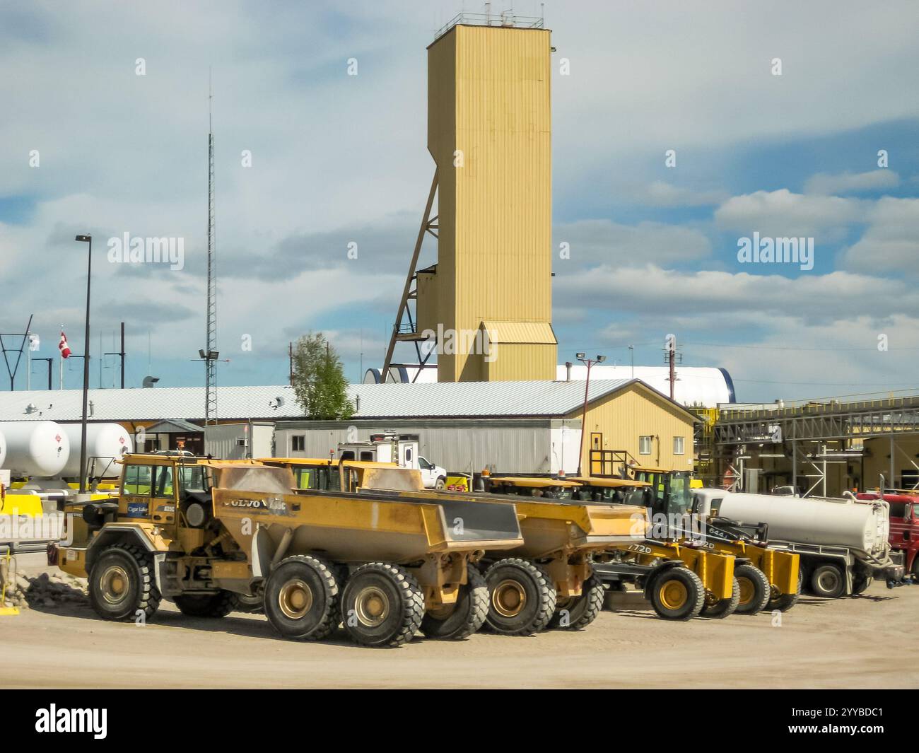 Un groupe de véhicules de construction sont garés dans un lot à côté d'un grand bâtiment. Les véhicules comprennent un camion à benne basculante, un tracteur et un camion. La scène est Banque D'Images