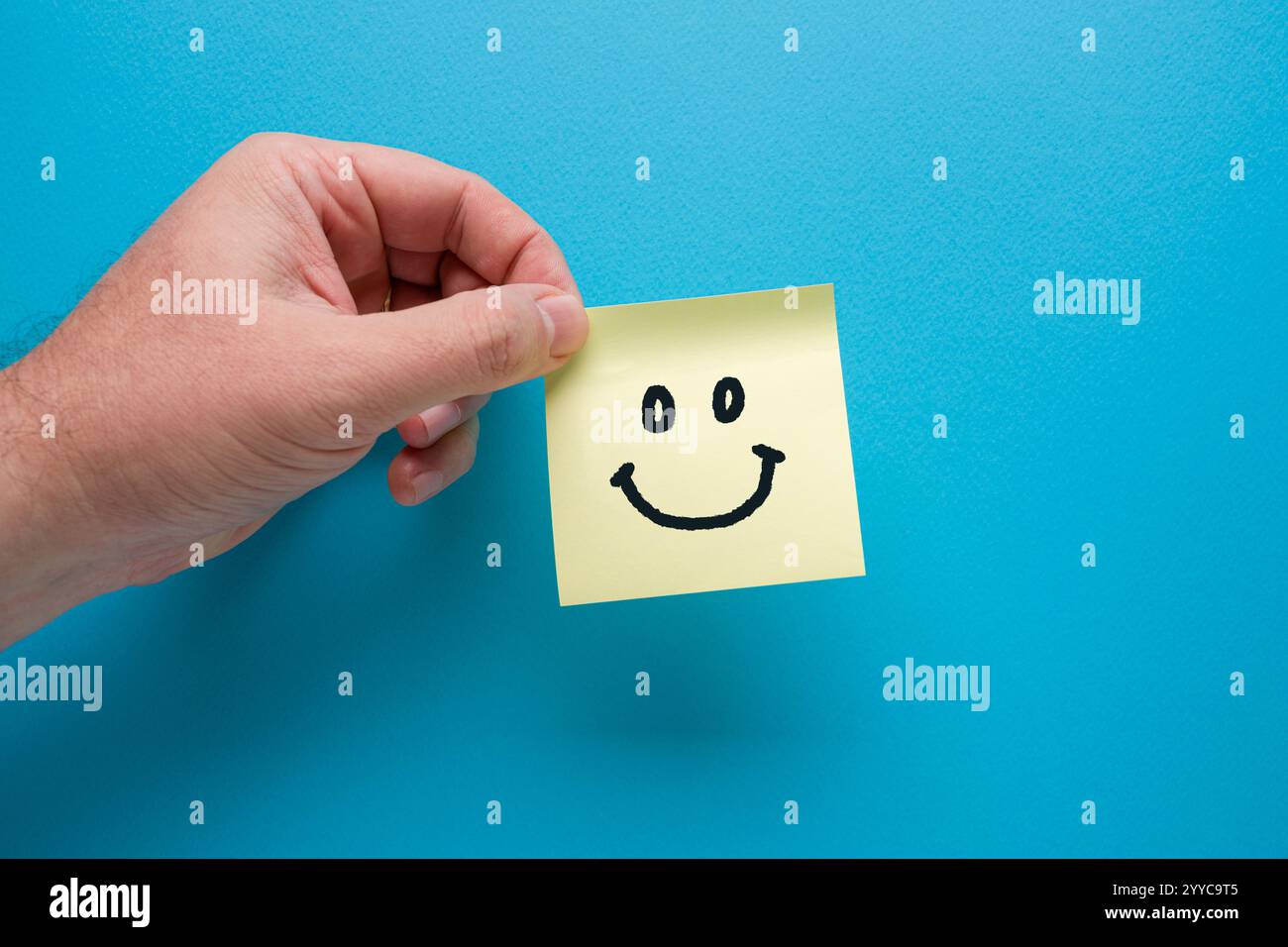 Main tenant du papier de note autocollante jaune ou post-it avec icône smiley sur fond bleu Banque D'Images