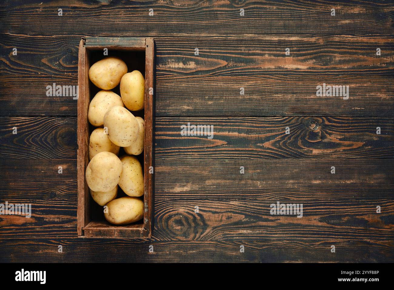 Une boîte en bois rustique remplie de pommes de terre fraîchement récoltées, reposant sur une table en bois sombre et texturée. Les couleurs naturelles et les matériaux soulignent le terreux Banque D'Images