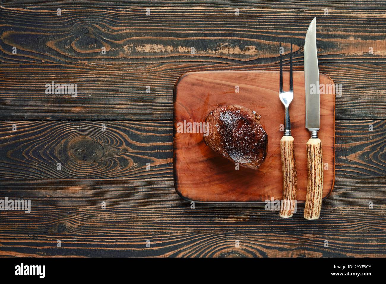 Un steak magnifiquement grillé repose sur une planche à découper rustique en bois, accompagné d'un couteau et d'une fourchette. Les tons chauds du bois contrastent avec la charcuterie Banque D'Images