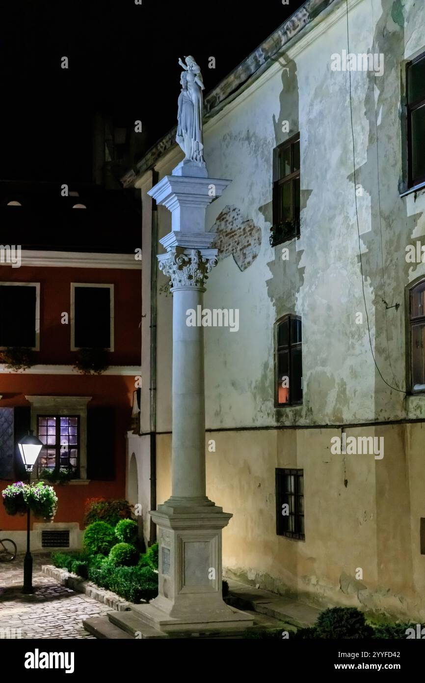 Une grande colonne blanche avec une statue d'une femme sur le dessus. Le bâtiment derrière la colonne est vieux et a beaucoup de fenêtres Banque D'Images