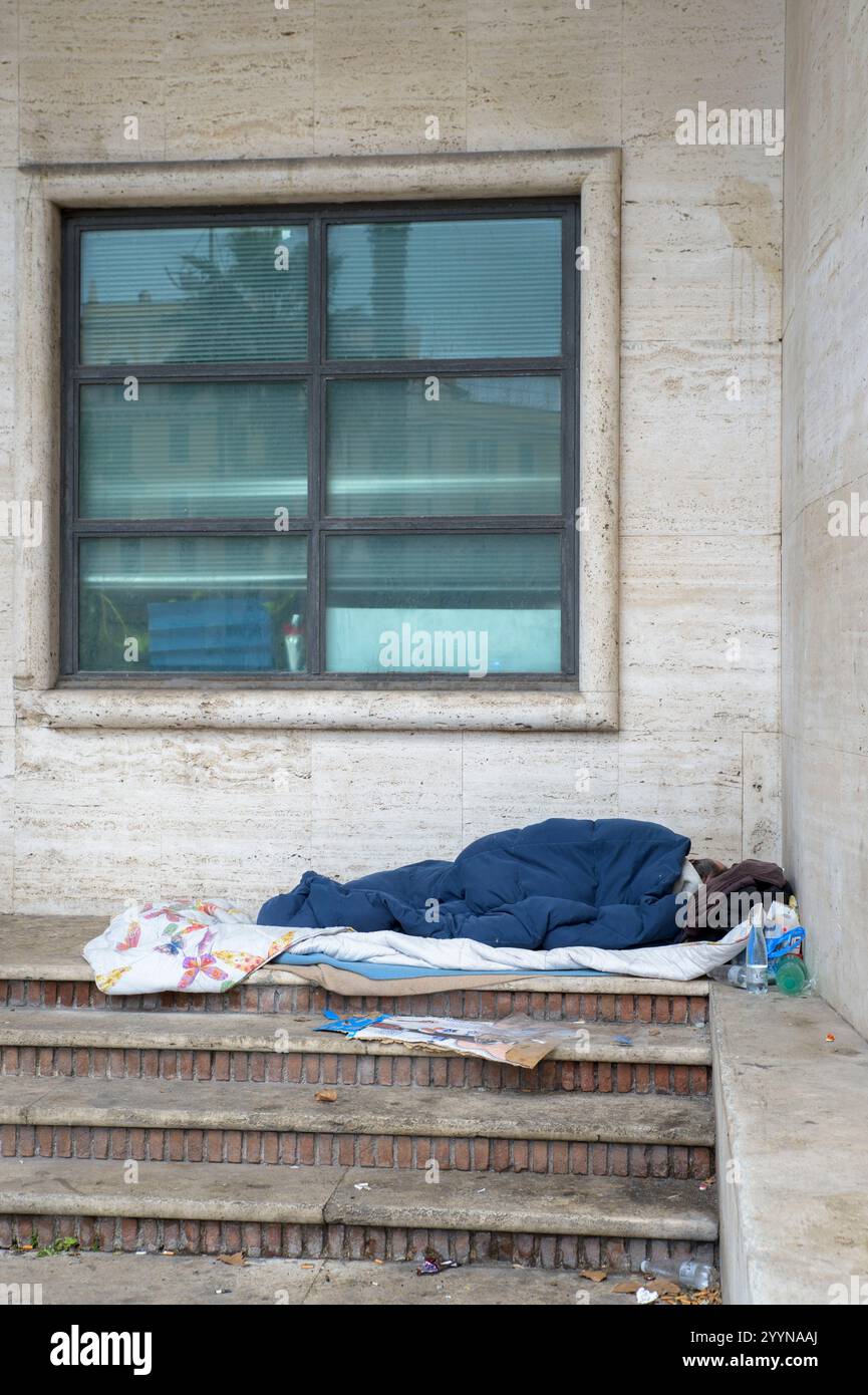 Rome, Italie. 22 décembre 2024. Un sans-abri dort dans la chaleur de son lit sous un auvent à l'abri du vent et de la pluie, tandis que le bâtiment en face se reflète dans le verre au-dessus, tard un dimanche matin de décembre à Rome. Selon le rapport du marché immobilier sur les ventes notariées et les prêts hypothécaires, publié le 19 décembre 2024 par l'Institut National de la Statistique (ISTAT), ''au premier trimestre 2024, après une tendance sensiblement stable observée au quatrième trimestre 2023, les transactions immobilières ont repris la tendance négative observée à partir du troisième trimestre 2022. T Banque D'Images