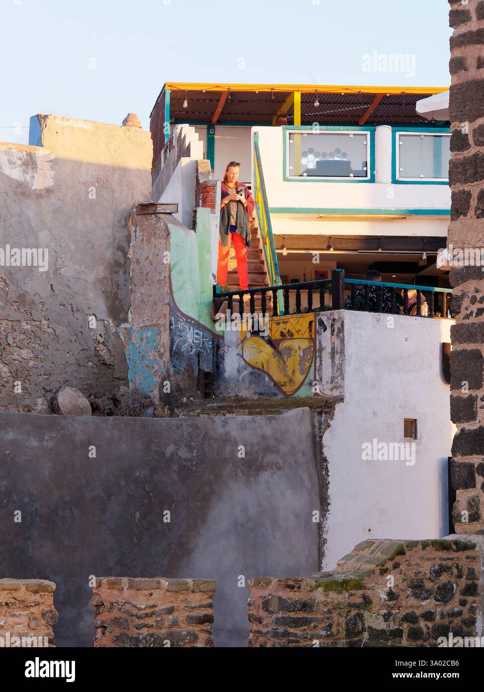 Femme en rouge quitte un petit restaurant rustique de base avec des rampes construites dans les murs de la ville un soir dans la médina à Essaouria, Maroc. Février/mars 2025 Banque D'Images