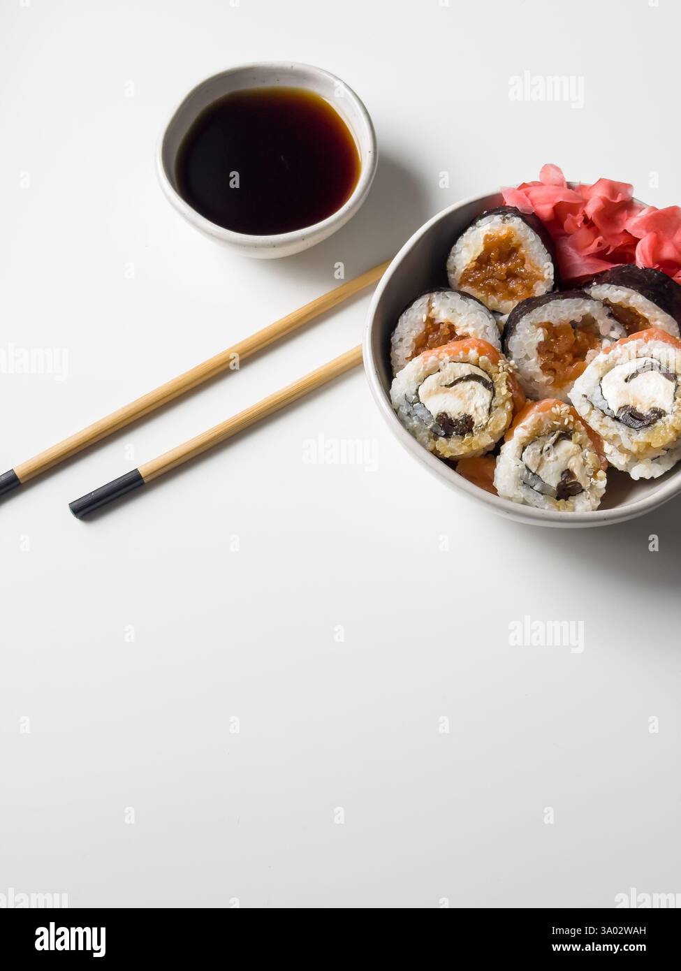 Rouleaux de sushi dans un bol avec du gingembre rose et de la sauce soja et des bâtonnets en bois sur la vue de dessus de table blanche Banque D'Images