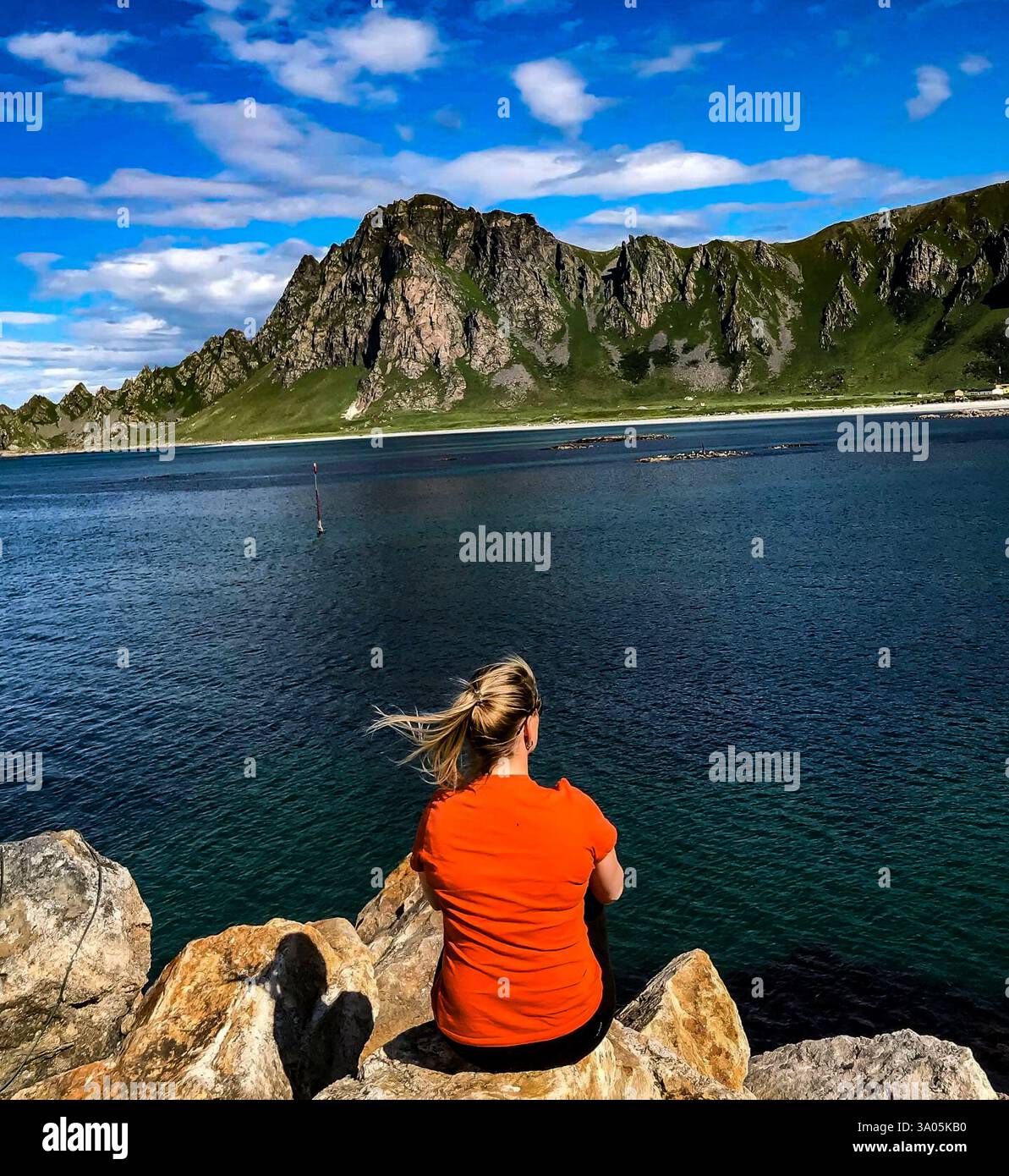 Une femme est assise sur les rochers côtiers, contemplant la mer turquoise et les montagnes escarpées de Bleik, Andøya, Nord-Norge. Un moment paisible. Banque D'Images