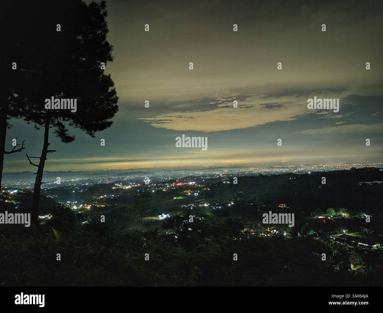 Une vue panoramique d'un paysage urbain illuminé sous un ciel nuageux spectaculaire la nuit. Le premier plan comprend des arbres et des terrains vallonnés, ajoutant de la profondeur au tra Banque D'Images