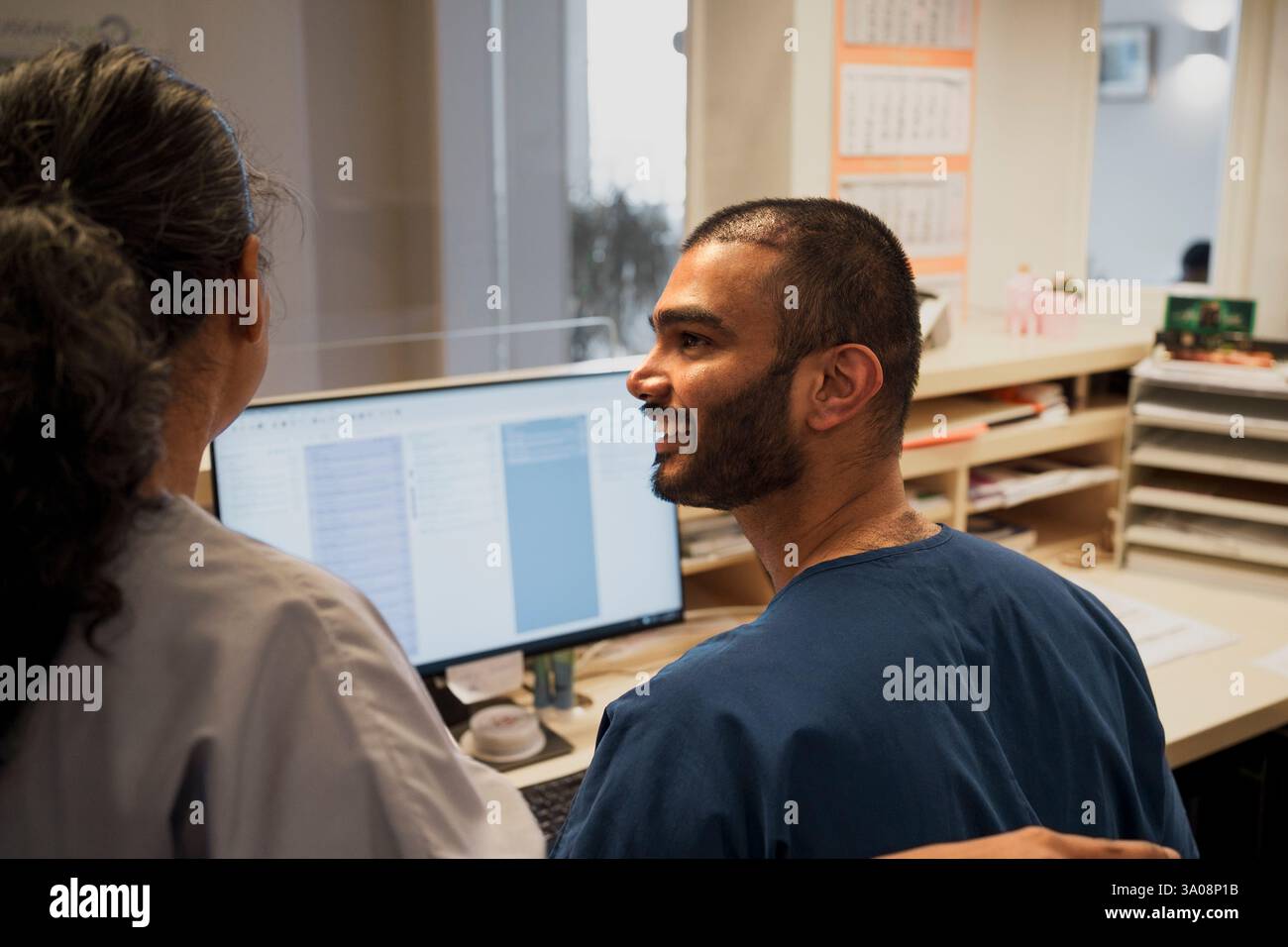 Médecin masculin souriant parlant avec une collègue tout en travaillant sur un PC de bureau à l'hôpital Banque D'Images