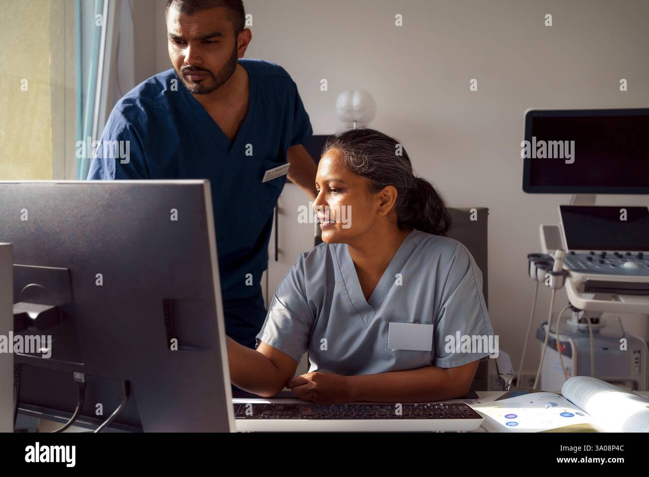 Professionnel de santé masculin debout près d'une collègue féminine utilisant un PC de bureau et discutant à l'hôpital Banque D'Images