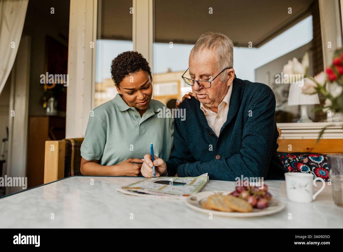 Homme senior résolvant le puzzle de mots croisés dans le journal tout en étant assis à côté de l'infirmière féminine à l'extérieur de la maison Banque D'Images