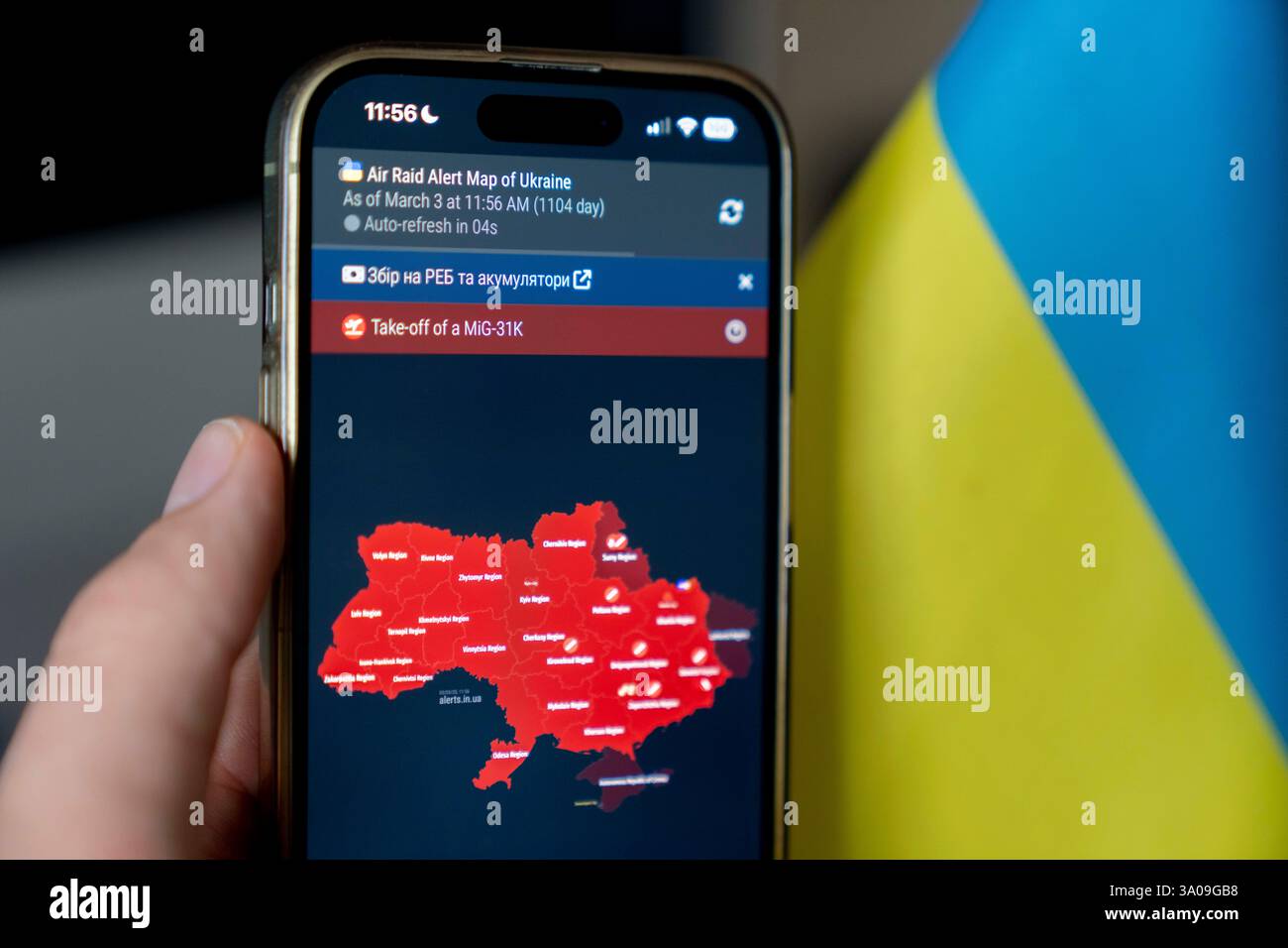 Illustration d'une personne tenant un smartphone en main à côté d'un drapeau ukrainien affichant une carte de l'Ukraine, marquant en rouge les régions menacées par la Russie, alors que des sirènes de raid aérien sonnent à travers Kiev. Une alerte MiG-31K a été déclarée dans toute l'Ukraine au moment de la prise de la photo. Banque D'Images
