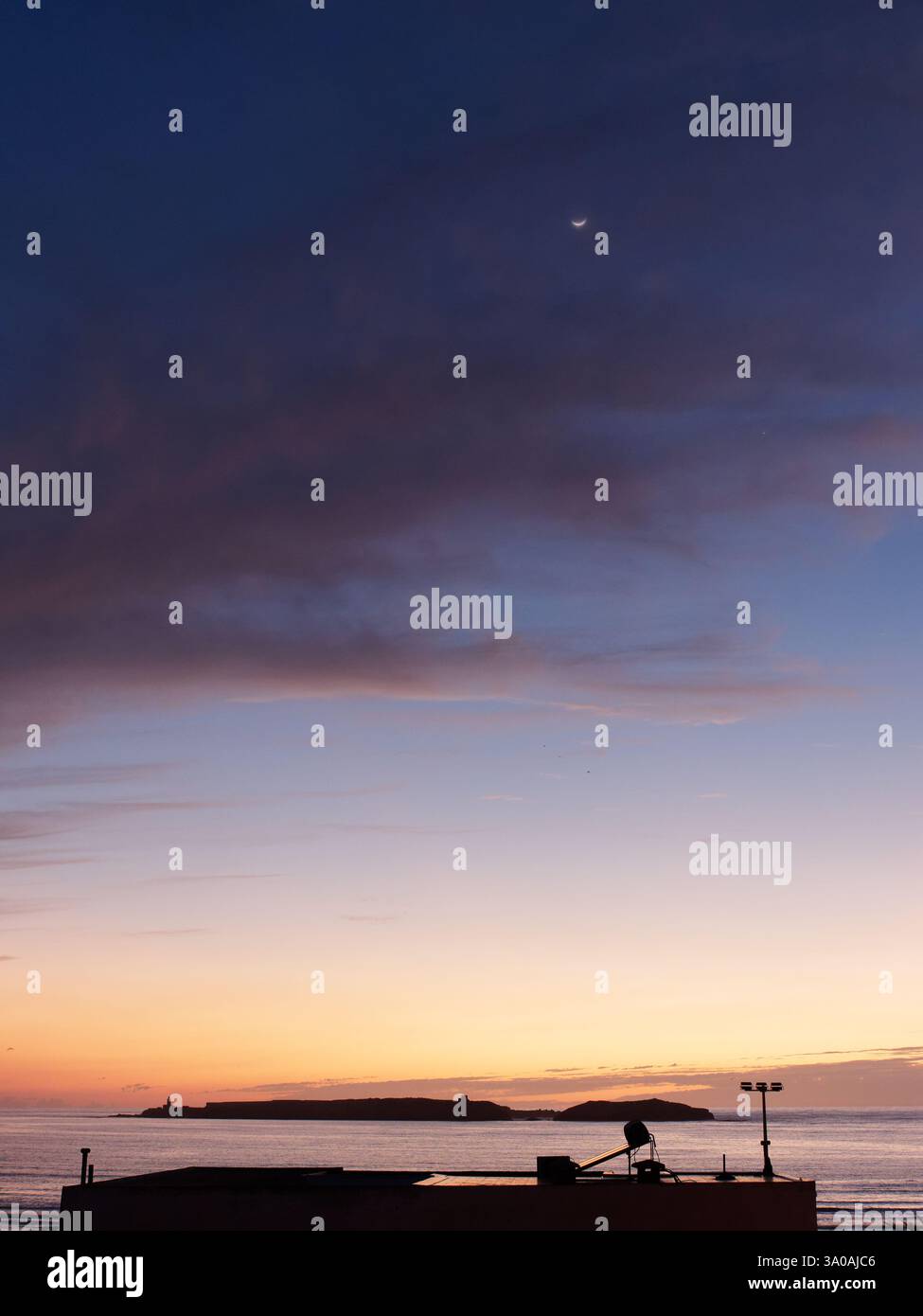 Lune dans le ciel sombre au coucher du soleil avec ciel rouge et île ci-dessous sur la mer Atlantique avec toit en premier plan à Essaouira. 2 mars 2025 Banque D'Images