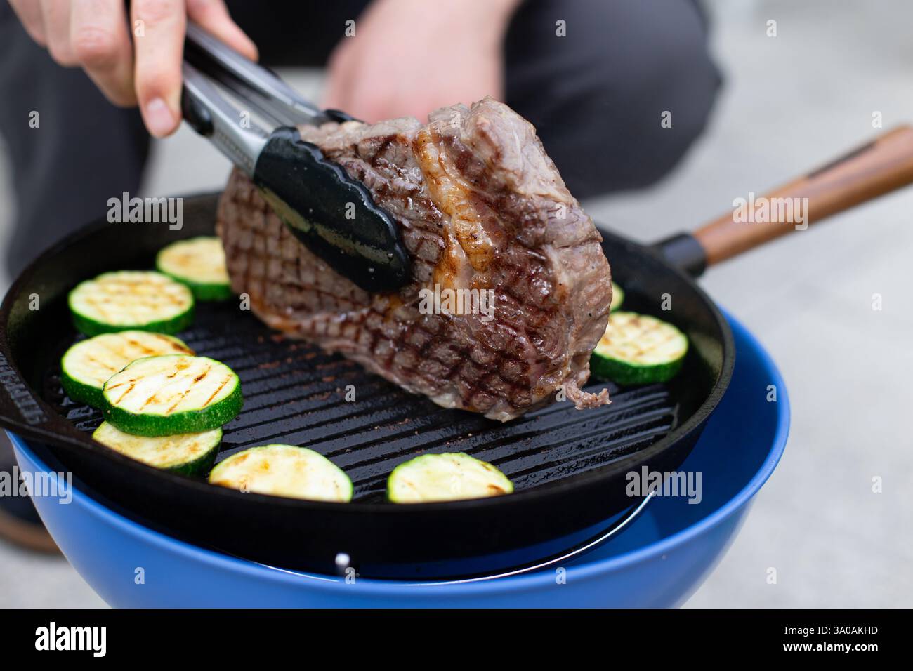 Un steak grillé juteux étant retourné avec des pinces sur une poêle en fonte, accompagné de courgettes tranchées. Une scène de cuisine extérieure parfaite pleine de saveur et Banque D'Images