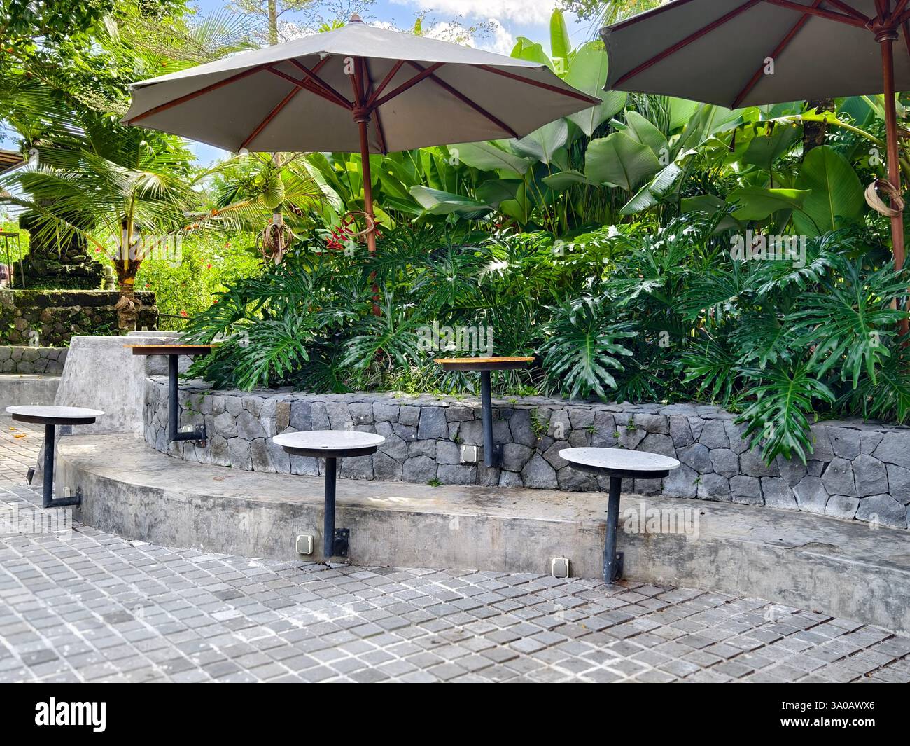 Un coin salon extérieur serein avec des tables rondes en pierre, construit dans des tabourets, et de grands parasols dans un café de verdure tropicale Banque D'Images