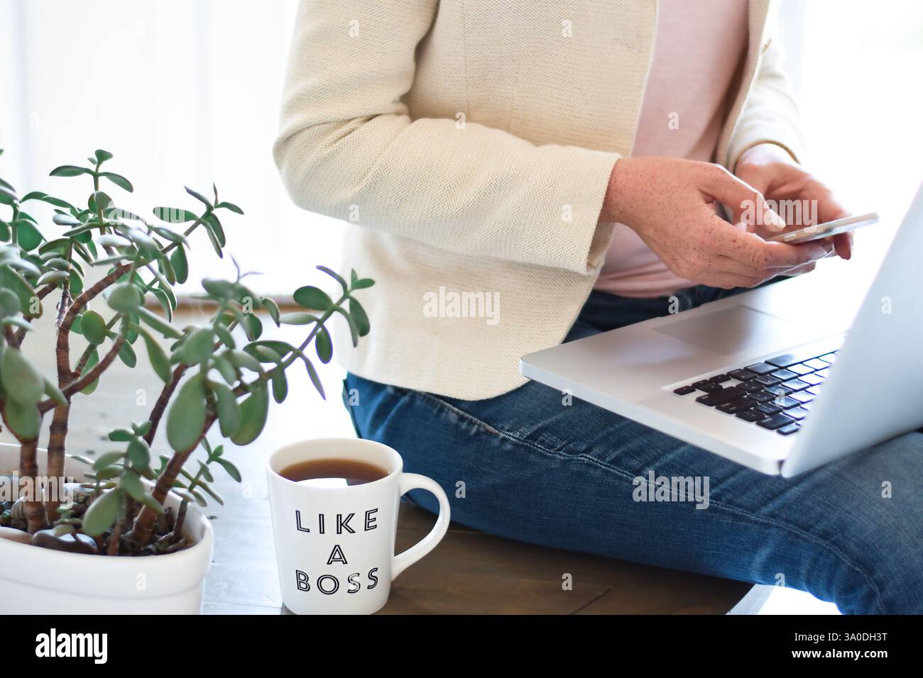 Femme assise sur le dessus du bureau avec un ordinateur portable utilisant un téléphone portable Banque D'Images Femme assise sur le dessus du bureau avec un ordinateur portable utilisant un téléphone portable Banque D'Images