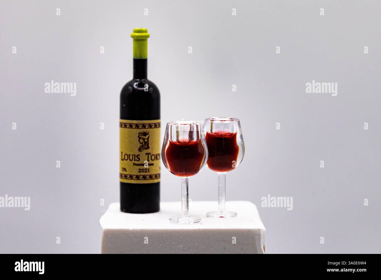 Une bouteille de vin miniature avec deux verres remplis sur un piédestal blanc. Cette photo macro capture les moindres détails, créant l'illusion d'un vin réel Banque D'Images