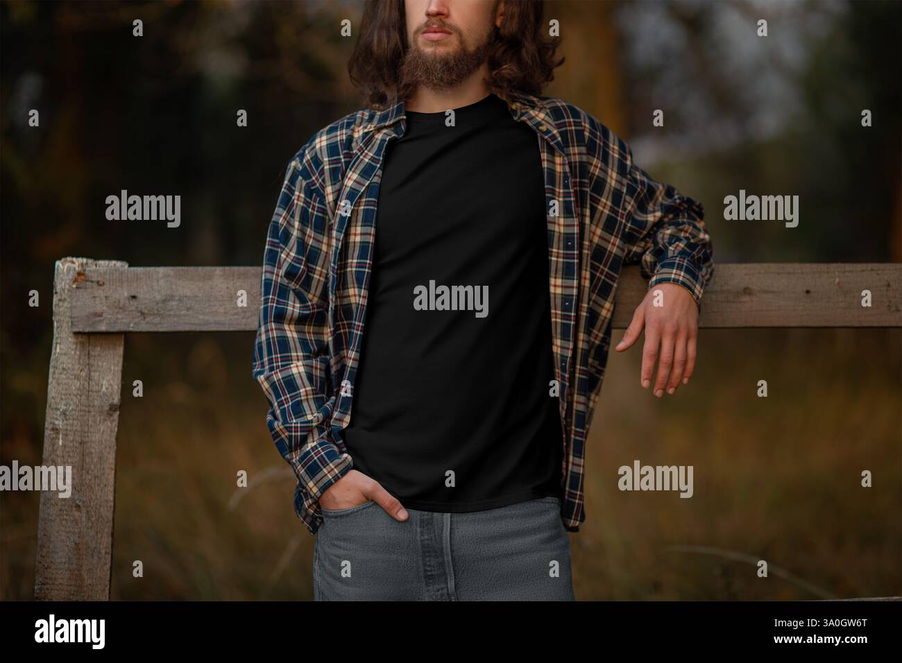 Modèle de t-shirt noir sur un gars avec une longue barbe, cheveux, dans une chemise en flanelle, appuyé contre une clôture de village, à l'extérieur, lumière du coucher du soleil, vue de face. Mâle c Banque D'Images