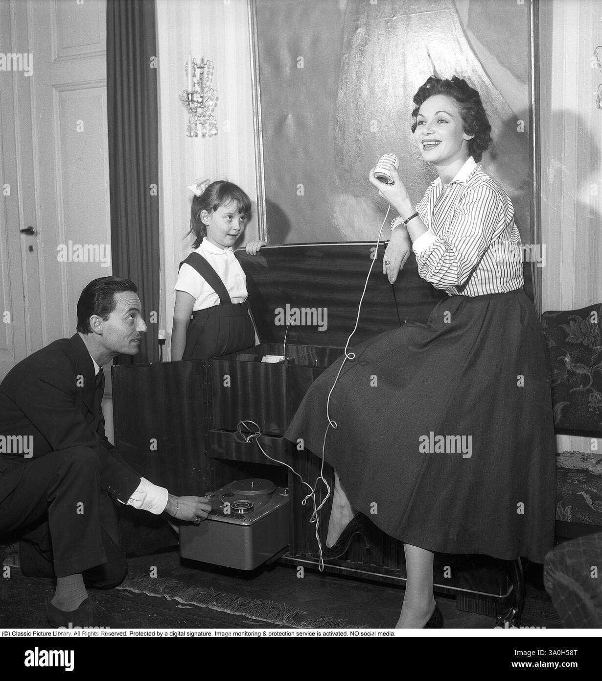 Famille à la maison 1956. Une scène domestique mettant en scène une famille avec un tourne-disque. Elle chante dans un micro au son de la musique. Il gère les paramètres du tourne-disques et de l'enregistreur. Une atmosphère joyeuse à un moment de divertissement et de convivialité. Se est la chanteuse actrice Ulla Sallert (1918-1986) avec son mari Franz von lampe et leur fille Thérèse von lampe. Kristoffersson réf. BU22-12 Banque D'Images