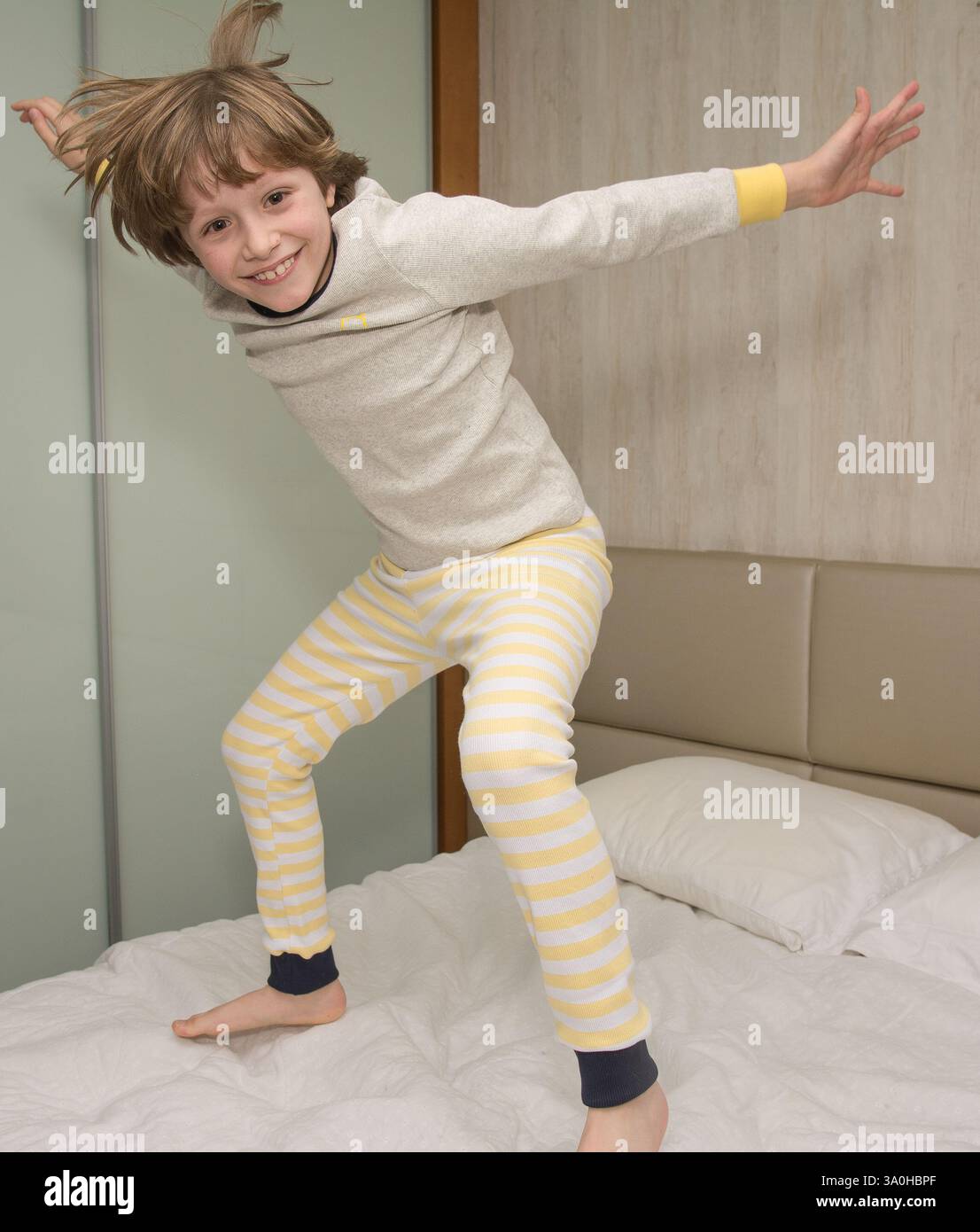 Un garçon joyeux avec un pyjama jaune rayé saute sur le lit, plein d'excitation et d'espiègle. Parfait pour la mode enfant, le plaisir du coucher et la maison Banque D'Images