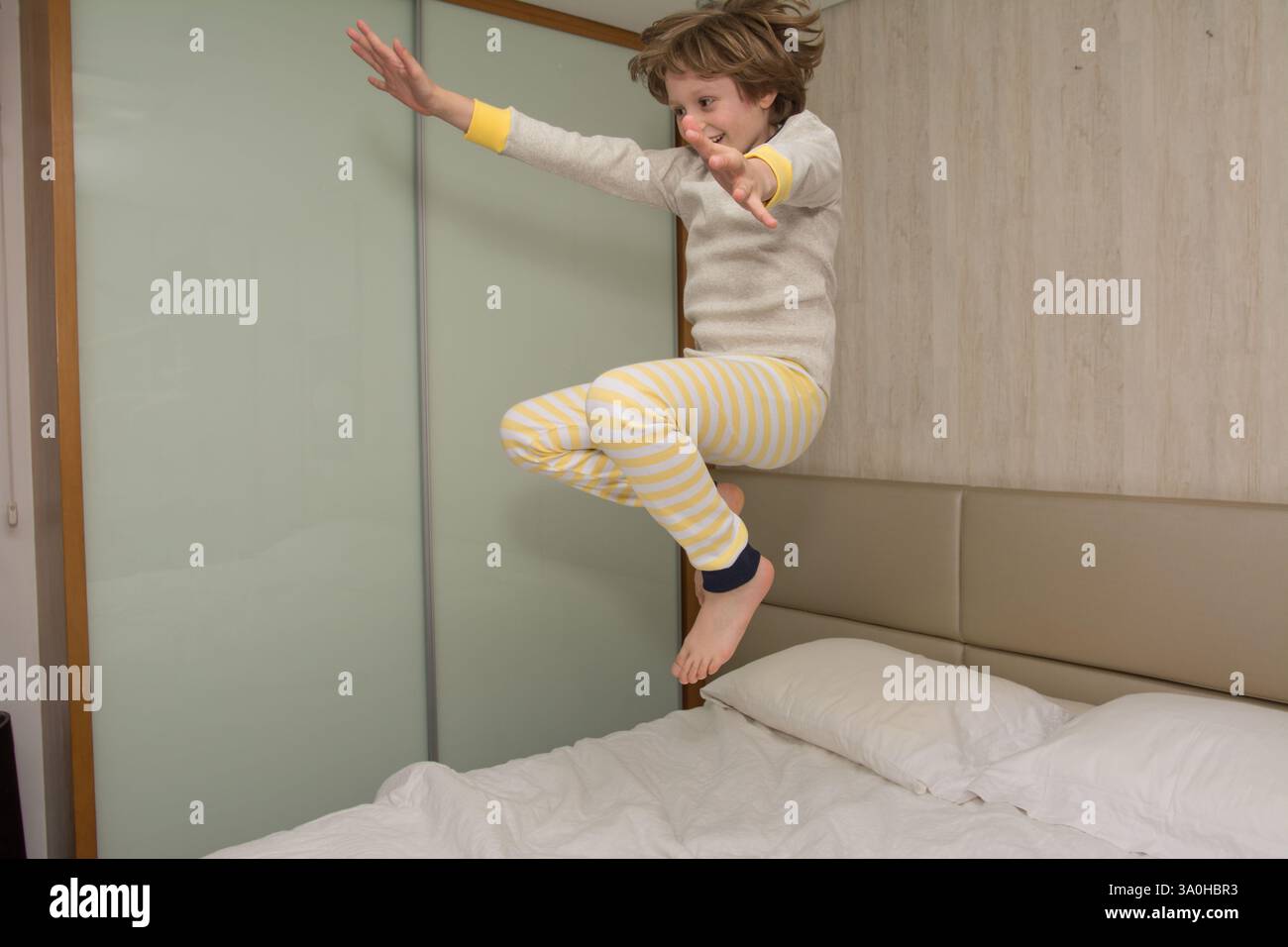 Un garçon joyeux en pyjama jaune rayé saute en plein air sur un lit confortable, capturant le plaisir insouciant de l'enfance. Parfait pour la mode des enfants, le jeu en intérieur, et ha Banque D'Images
