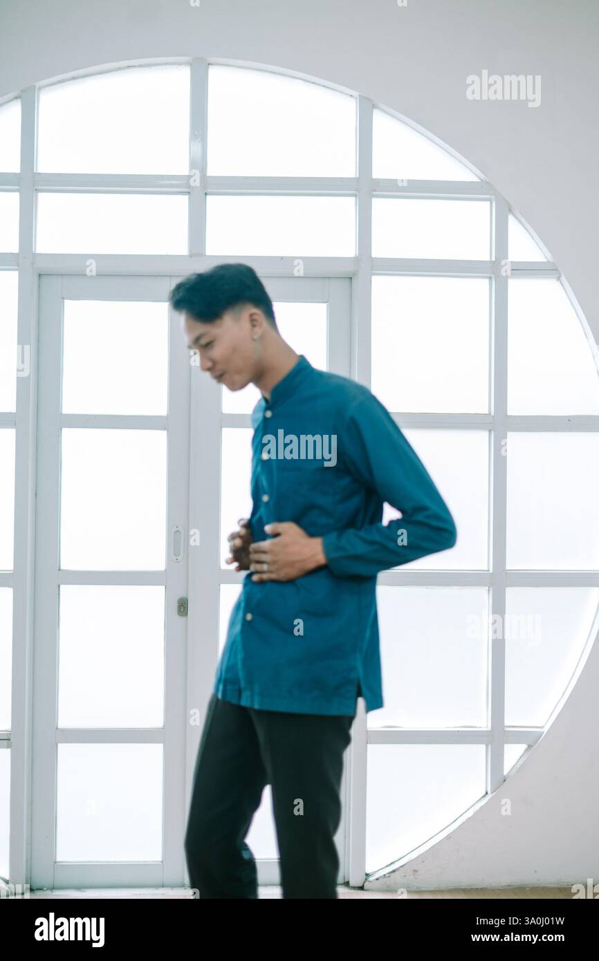 Un jeune homme contemplatif dans une chemise bleue et un pantalon noir tient soigneusement son abdomen. Posant près d'une fenêtre blanche brillante, il respire le style moderne an Banque D'Images