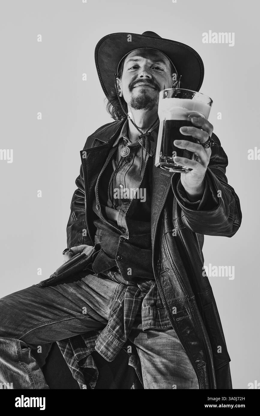 Portrait noir et blanc d'un bel homme souriant en manteau de cuir, jeans et chemise portés, tenant un verre de bière mousseuse sombre, appréciant la boisson Banque D'Images
