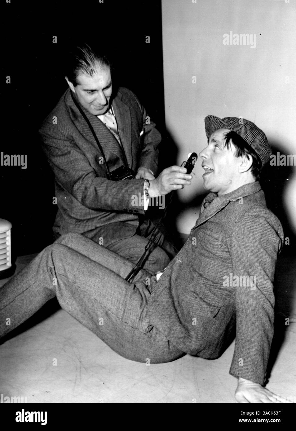 Le photographe et le comédien Norman Wisdom, le comédien de 38 ans qui vient de prouver qu'il peut être aussi drôle sur les films qu'à la télévision britannique, hier veulent voir Baron, le photographe. Ici Baron (agenouillé) essaie d'obtenir un gros plan de sagesse qui est vêtu dans ses vêtements de scène d'un petit costume gris serré et d'une casquette en tissu. 16 décembre 1953. Banque D'Images