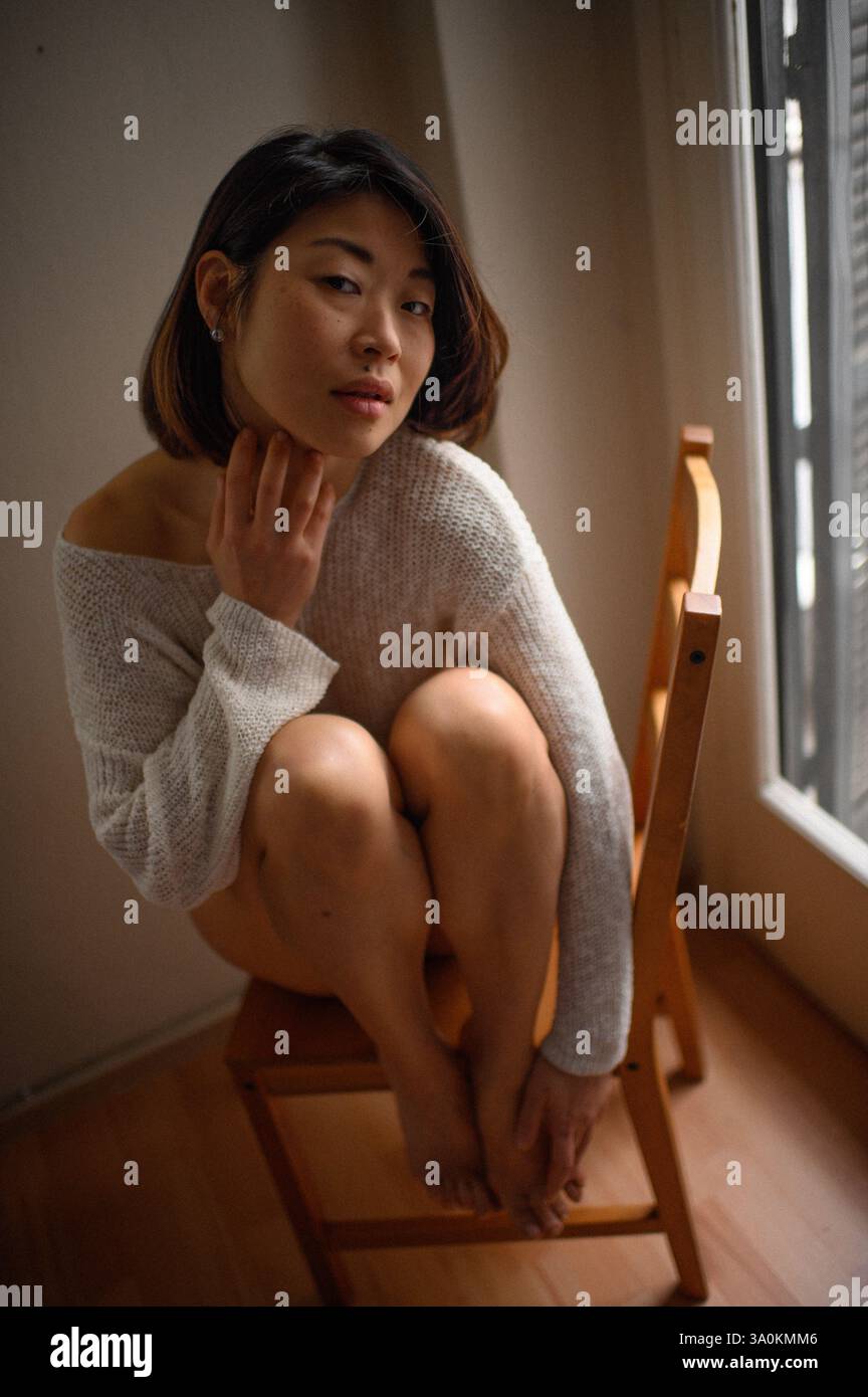 Femme asiatique aux cheveux courts assise sur une chaise en bois près de la fenêtre à la maison, profitant d'un moment de calme à l'intérieur. Banque D'Images