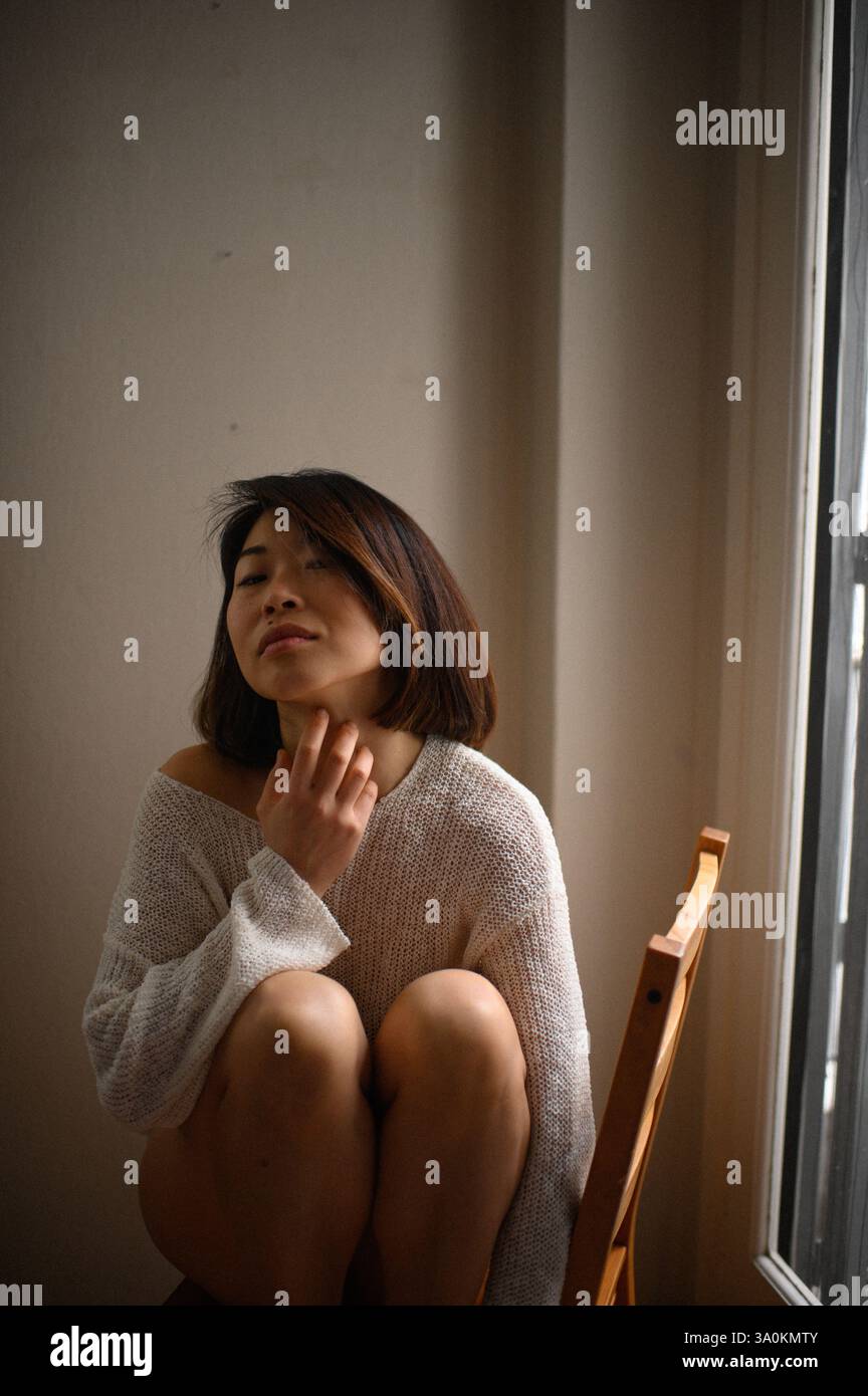 Femme asiatique aux cheveux courts assise sur une chaise en bois près de la fenêtre à la maison, profitant d'un moment de calme à l'intérieur. Banque D'Images