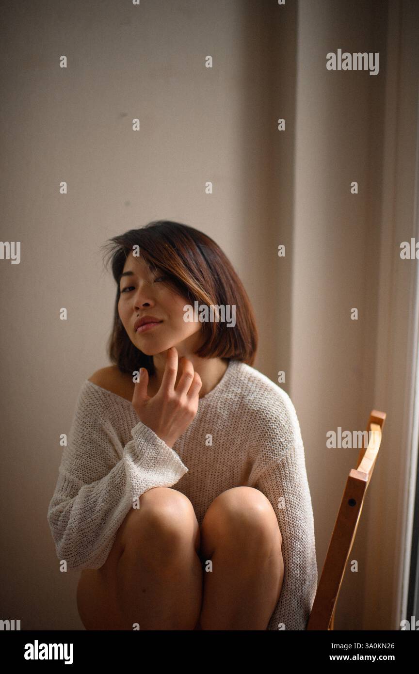 Femme asiatique aux cheveux courts assise sur une chaise en bois près de la fenêtre à la maison, profitant d'un moment de calme à l'intérieur. Banque D'Images