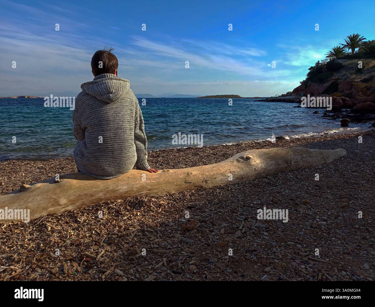 Athènes, Grèce- mars 2, 2025 - Une figure féminine est assise sur une bûche de bois flotté altérée, face à la mer sans fin. Banque D'Images