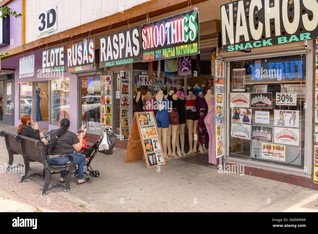 Deux femmes sont assises sur un banc de trottoir, enfant dans une poussette, devant le Snack Bar nachos et magasin de vêtements avec mannequins devant, McAllen, Texas, États-Unis. Banque D'Images