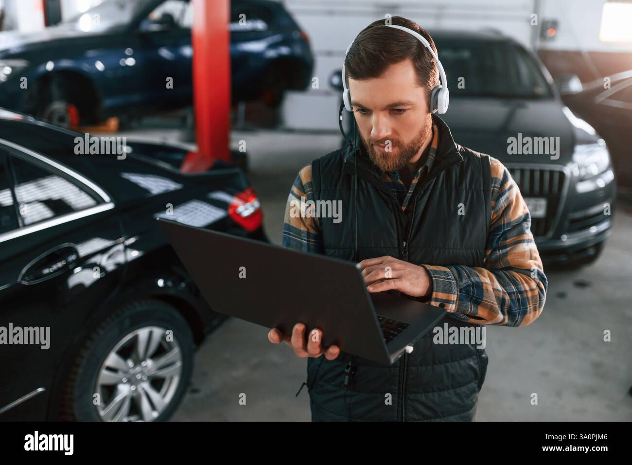 Debout avec l'ordinateur portable dans les mains et dans les écouteurs. L'homme en uniforme travaille dans le salon automobile. Banque D'Images