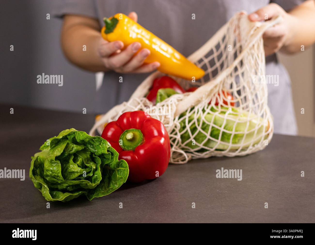 Gros plan de mains féminines qui placent des légumes frais de saison dans un sac ECO net sur une table de cuisine grise Banque D'Images