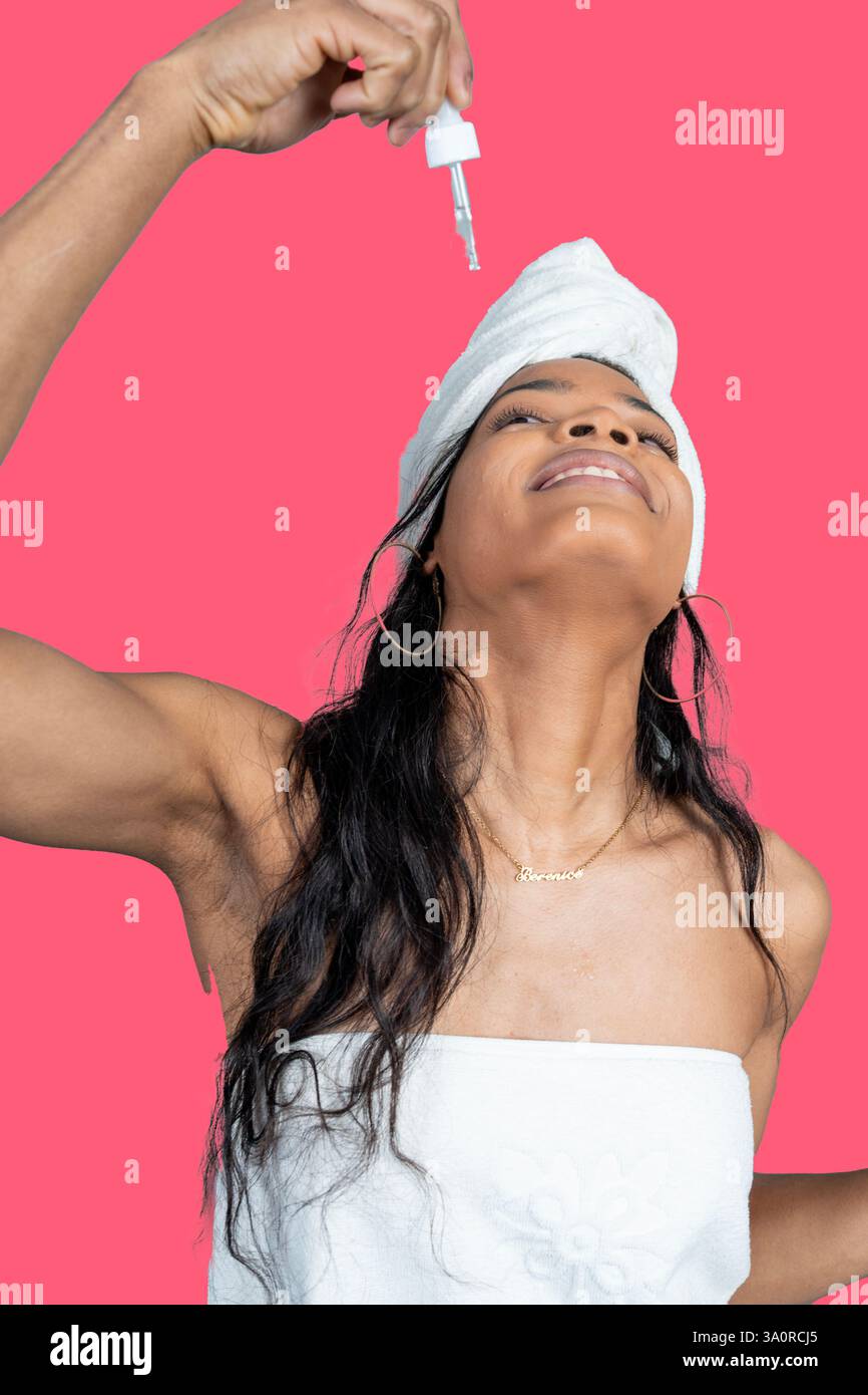 Femme souriante portant une serviette appliquant le sérum sur le visage avec compte-gouttes après la douche, sur fond rose Banque D'Images