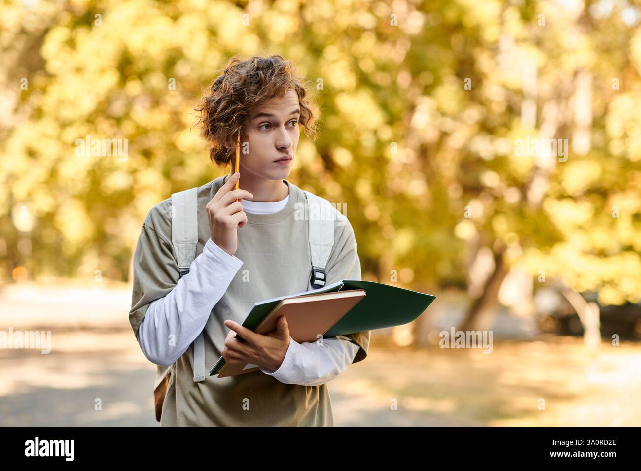 Un jeune homme aux cheveux élégants tient des cahiers et un crayon, profondément dans la pensée parmi les arbres dorés. Banque D'Images