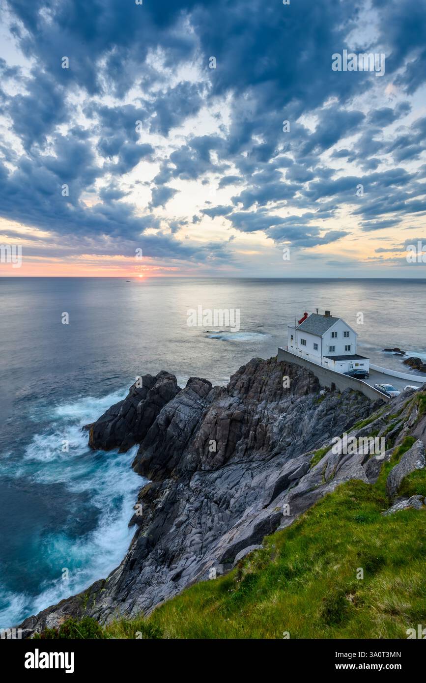 Un magnifique coucher de soleil projette des couleurs vives sur la côte norvégienne, mettant en évidence une maison blanche sur des falaises rocheuses au milieu de vagues écrasantes et de nuages spectaculaires Banque D'Images