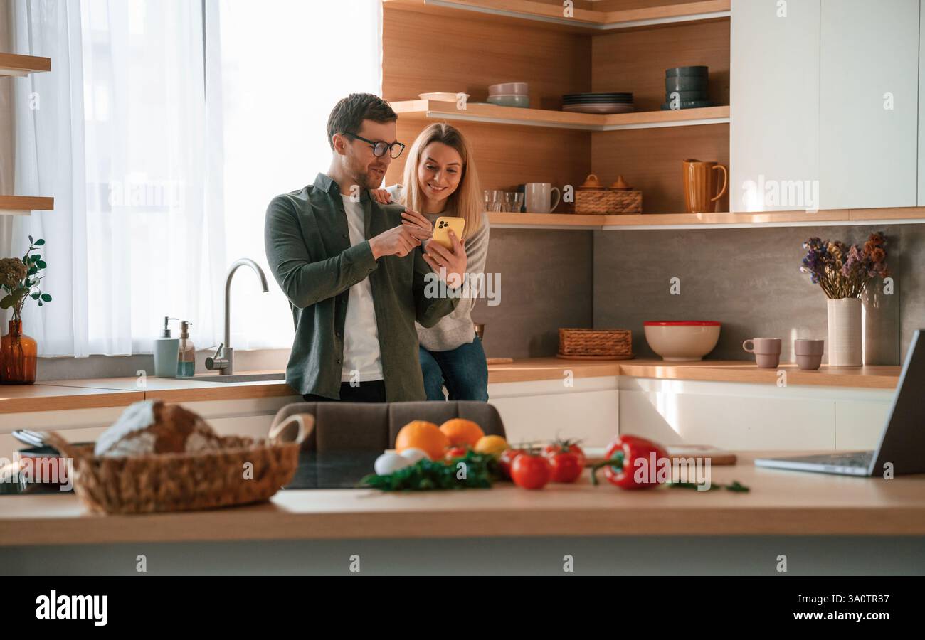 Homme montrant des médias à la femme au téléphone. Jeune couple est sur la cuisine ensemble. Banque D'Images