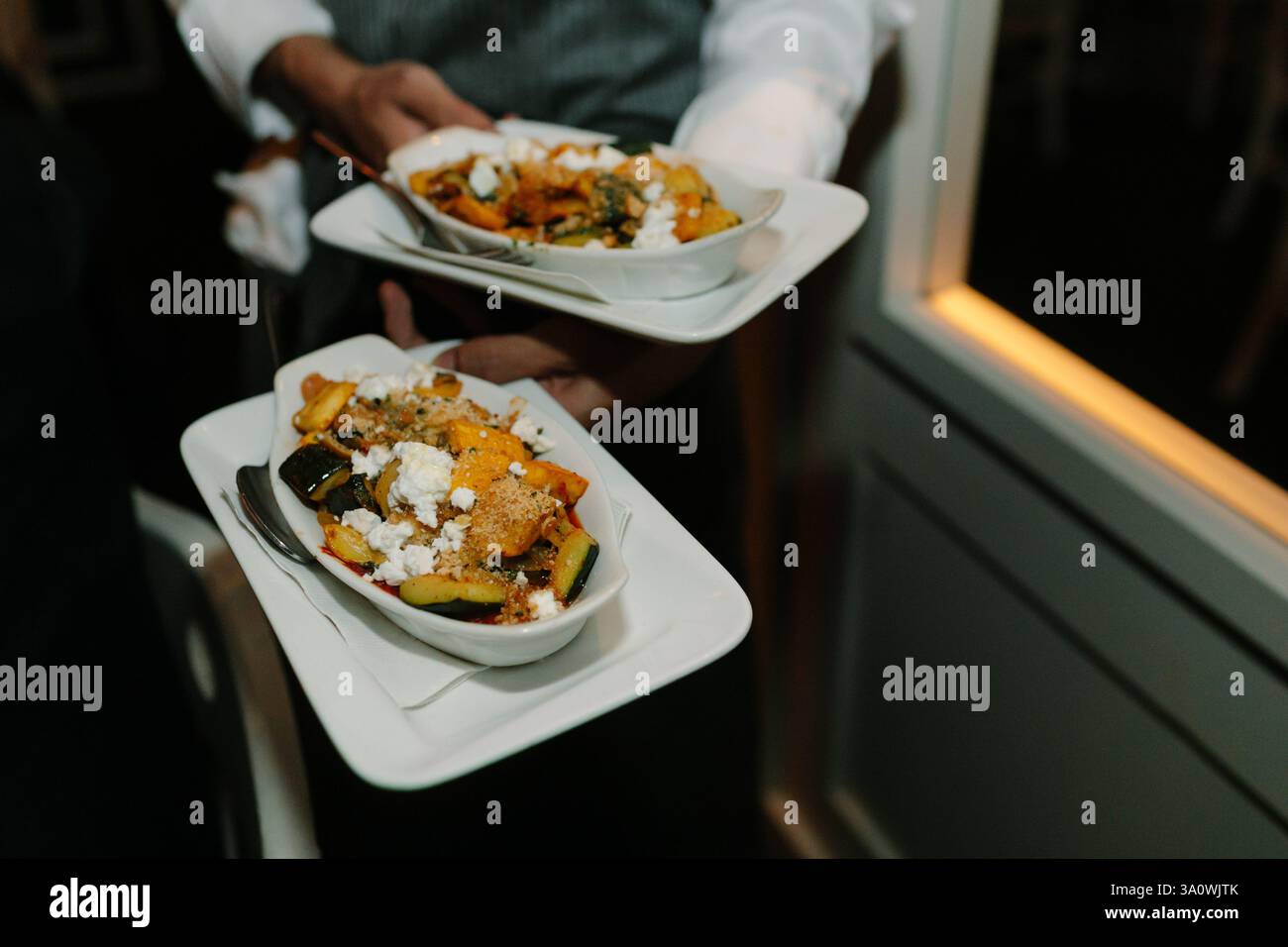 Un serveur sert deux plats magnifiquement plaqués, mettant en valeur des légumes et des garnitures vibrants, dans un restaurant faiblement éclairé. Banque D'Images