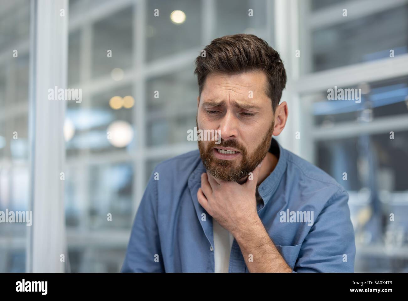 Un homme inquiet vêtu d'une chemise bleue décontractée tient son cou exprimant l'inconfort et la maladie, souffrant peut-être d'un mal de gorge. Banque D'Images