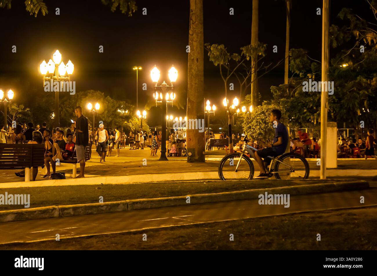 Vue de la place Menino João Helio zone piétonne animée avec des personnes profitant de bancs, de vélos et d'interactions sociales sous les lumières lumineuses de la nuit d'été. Banque D'Images