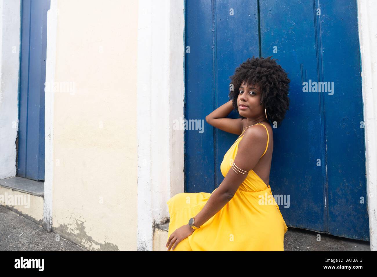 Belle jeune femme aux cheveux afro, vêtue d'une robe jaune, assise près d'une porte bleue d'une vieille maison. Personne confiante. Brésil Banque D'Images