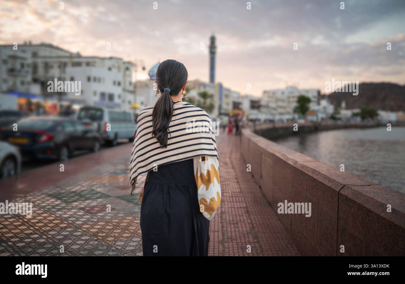 Femme en tenue décontractée marche le long de la corniche de Muttrah à Muscat, Oman, immergée dans la vue panoramique sur le front de mer avec la lumière de l'heure d'or montrant le cha Banque D'Images